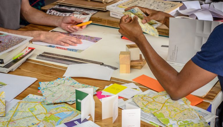 A table is covered in old maps, glue and pieces of paper that are being used to make something.